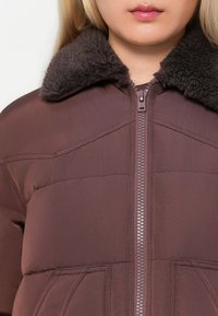 Giubbotto imbottito marrone con zip e colletto in pelliccia scura. Presenta sezioni trapuntate e due tasche frontali. Texture esterna liscia.