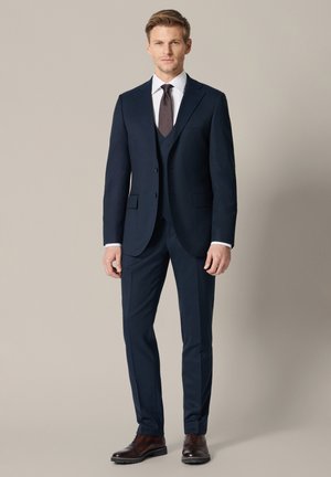 Młody mężczyzna stoi, ubrany w granatowy garnitur trzyczęściowy, białą koszulę, ciemny krawat i brązowe skórzane eleganckie buty na jednolitym tle.
