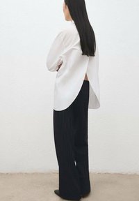 Wit shirt met lange mouwen en een gespleten rug, gecombineerd met zwarte pantalon met krijtstreep en zwarte puntige schoenen, staand tegen een effen achtergrond.