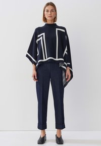 Marineblauer Poncho mit weißen geometrischen Mustern, kombiniert mit dunklen Jeanshosen, die an den Bündchen umgeschlagen sind. Schwarze Slipper runden den Look ab.