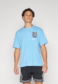 Világoskék pamut póló, kék és sárga négyzet alakú grafikával, Nike logóval a bal mellen. Rövid ujjak és kerek nyakkivágás.