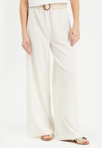 Pantalons à jambes larges en crème avec un tissu texturé, une ceinture tressée avec une boucle ronde, associés à des sandales plates.