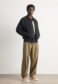 Young man standing with hands in pockets wearing black bomber jacket, white shirt, tan wide-leg pants, and beige sneakers.