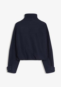 Laivastonsininen lyhyt takki, jossa on korkea kaulus ja pitkät hihat säädettävillä napillisisilla kalvosimilla, kuvattuna takaa.