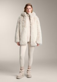 Kabát z umelé kožušiny svetlo béžovej farby s kapucňou, doplnený krémovým rolákom, béžovými legínami a členkovými topánkami s textúrou. Mäkký, objemný dizajn.