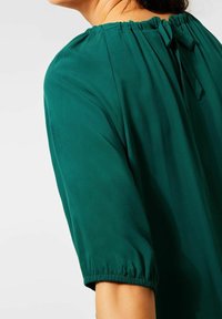 Blusa verde realizzata in tessuto morbido, con scollatura arricciata e maniche corte a palloncino con polsini elasticizzati. Si lega sul retro.