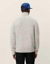 Hellgrauer Strickpullover mit hohem Kragen, geripptem Saum und gesprenkelter Textur. Getragen mit einer blauen Mütze, die Text auf der Rückseite hat.