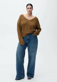 Bruin, geribd brei trui met een ruime V-hals, gecombineerd met hooggewaagde, losse blauwe jeans. Model draagt zwarte sandalen en een ronde ketting.