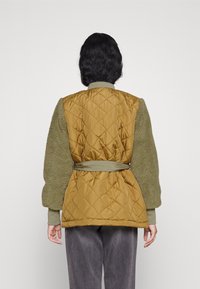 Quiltad bomberjacka i senapsgul med ärmar av grön konstpäls och bälte i midjan. Har ribbade muddarna och en enfärgad krage.