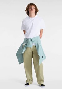 Vit t-shirt, mintgrön skjorta knuten i midjan, ljusbruna vida byxor och svarta slip-on skor med gula detaljer. Avslappnad outfitpresentation.