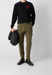 Pull noir avec logo "HUGO" rouge, pantalon vert olive, veste en cuir noire et chaussures noires, présentant une tenue décontractée moderne.