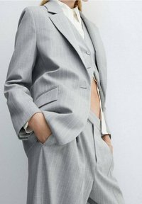 Harmaa viiruhaalari, jossa on muotoiltu bleiseri ja liivi. Kankaan pinta on sileä, ja bleiserissä sekä housuissa on taskut.