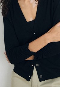 Svart cardigan med V-ringning och samlade ärmdetaljer, med knäppningar fram. Mjuk, stickad textur.