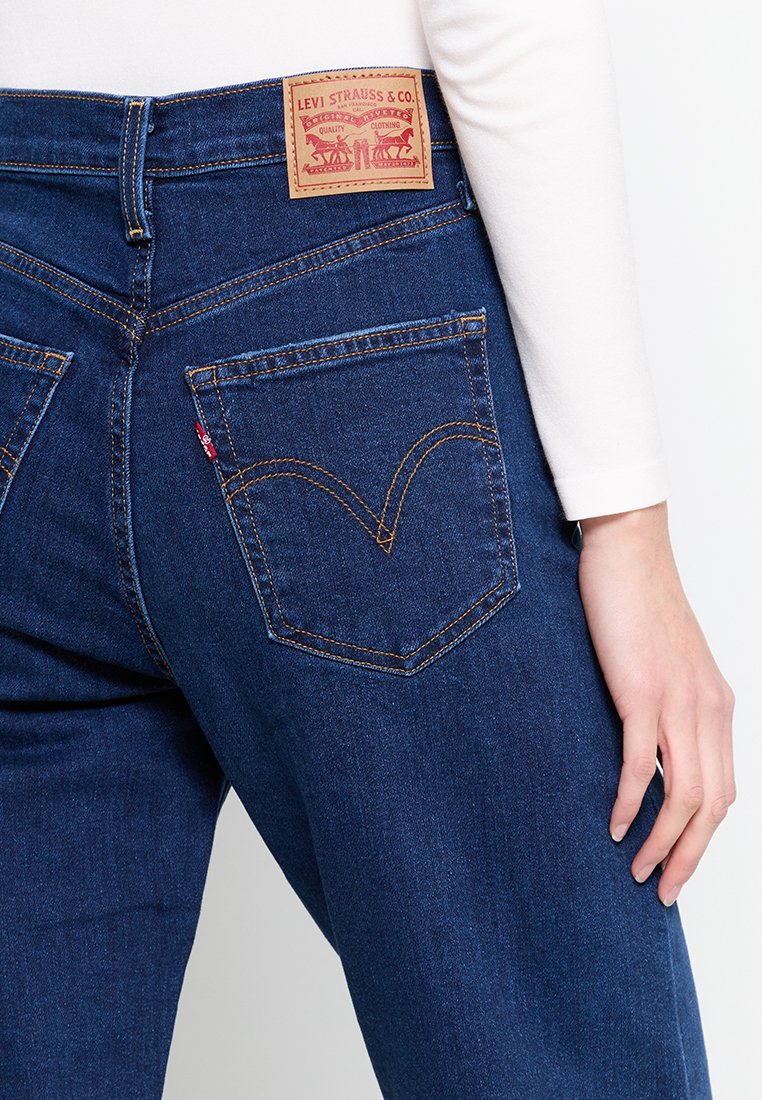 Jeans in denim blu scuro con etichetta in pelle marrone caratterizzata da un design rosso e oro. Include tasche posteriori distintive con un dettaglio di cucitura curvata.