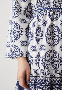 Rochie albă cu modele florale și geometrice albastre. Are mâneci lungi, talie strânsă și volane la margine. Fabricată dintr-un material ușor.