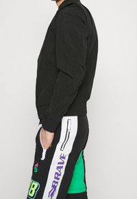 Černá bomberka vyrobená z texturované tkaniny, s předním zipem a žebrovanými manžetami. Nosená s kontrastními černo-zelenými kalhotami s vzorem.