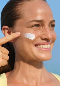 Krem nawilżający nakładany na policzek, ukazujący gładką, białą teksturę na opalonej skórze. Włosy są spięte do tyłu, odsłaniając czyste, neutralne tło.