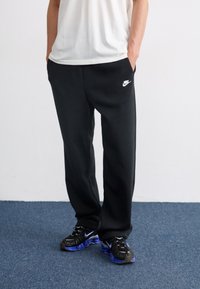 Črne trenirke z udobnim krojem, katerih posebnost so stranske žepe in bel Nike logotip na levem stegnu. V kombinaciji s črnimi in modrimi športnimi čevlji.