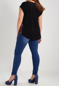 Sort kortærmet top med mønstret side, parret med blå skinny jeans og blå chunky-heeled sandaler, set bagfra.