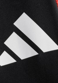 Schwarze Textiloberfläche mit einem auffälligen weißen Drei-Streifen-Logo-Design, glatte Textur mit matter Oberfläche, akzentuiert durch leuchtend rote Elemente.