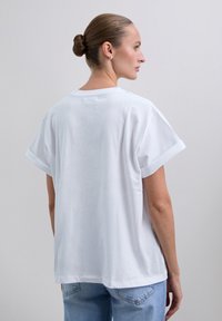 Donna con i capelli raccolti in uno chignon, indossa una semplice maglietta bianca a maniche corte e jeans azzurri chiari, girata di spalle davanti a uno sfondo uniforme.