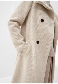 Cappotto beige a spina di pesce con un tessuto morbido e strutturato, design doppiopetto, bottoni neri e tasche laterali.