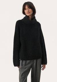 Zwarte gebreide trui met een hoge, omgeslagen kraag, geribbelde textuur en losse, lange mouwen. Gepaard met grijze op maat gemaakte broek.