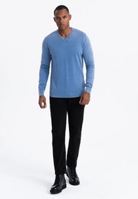 Suéter azul claro de cuello en V, textura de punto, combinado con pantalones negros y botas negras con cordones. El modelo se presenta con una expresión neutral.