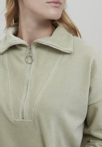 Ljusgrön kordflodzip-parka med hög krage, med en metallblixtlås och en geometrisk sömdetalj framtill. Mjuk textur.