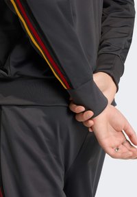 Zwarte sportjack met rode, gele en zwarte strepen op de mouwen. Heeft geribbelde boorden en een soepel textuur, gecombineerd met bijpassende broek.