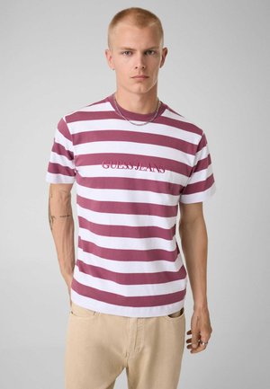 Młody mężczyzna z krótkimi blond włosami nosi bordowo-biały pasiasty t-shirt Guess Jeans i beżowe spodnie na jednolitym tle.