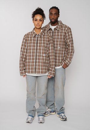 Mann und Frau stehen nebeneinander und tragen passende braun karierte Jacken, helle Jeans und Turnschuhe vor einem schlichten Hintergrund.