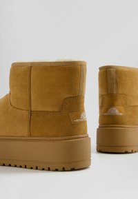 Tan suede ankle boots with a soft fur lining, stitched detailing, and a rubber sole featuring a tread pattern. Brand logo on the side.