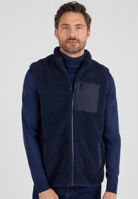Navyblauer Fleece-Weste mit einer Reißverschlusstasche, strukturiertem Obermaterial und glatten unteren Kanten. Verfügt über einen hohen Kragen und eine Frontreißverschluss.
