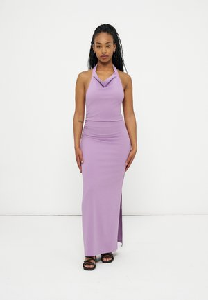 Woman standing wearing a sleeveless lilac halter-neck maxi dress with side slit and black strappy sandals against white background.
