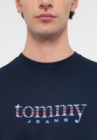 Tricou din bumbac bleumarin cu guler rotund striat, având un logo colorat "tommy" cu dungi roșii, albastre și violet, și cu "JEANS" dedesubt.