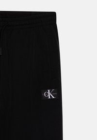 Black jogger pants with an elastic waistband, side pockets, and a CK logo patch on the thigh. Fabric has a smooth texture.