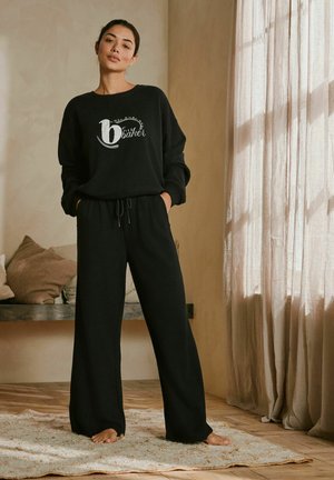 Melnā sporta jaka ar balto drukāto logo, pārī ar platām melnām biksēm, kas izgatavotas no teksturēta auduma, iekārtota mājīgā iekštelpās.