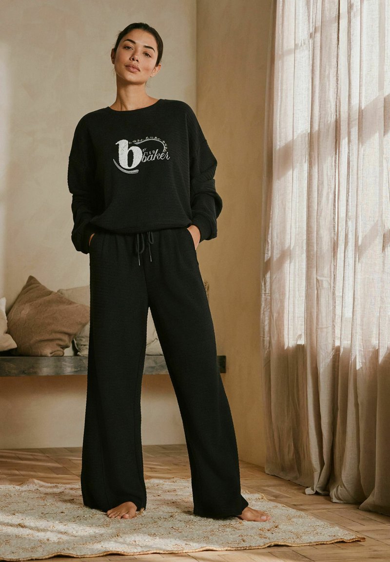 Schwarzer Sweatshirt mit einem weißen Logo, kombiniert mit weit geschnittenen schwarzen Hosen aus strukturiertem Stoff, in einer weichen, gemütlichen Innenumgebung.