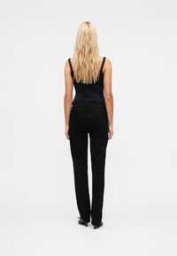 Ξανθιά γυναίκα με κυματιστά μαλλιά στέκεται με την πλάτη, φορώντας μαύρο αμάνικο τοπ, μαύρο παντελόνι κανονικής γραμμής και μαύρες ψηλοτάκουνες γόβες.