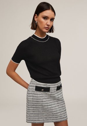 Schwarzes Strickoberteil mit kurzen Ärmeln, kontrastiert durch weiße Verzierungen. Kombiniert mit einem gestreiften Minirock, der schwarze Flecken und goldene Akzente aufweist.