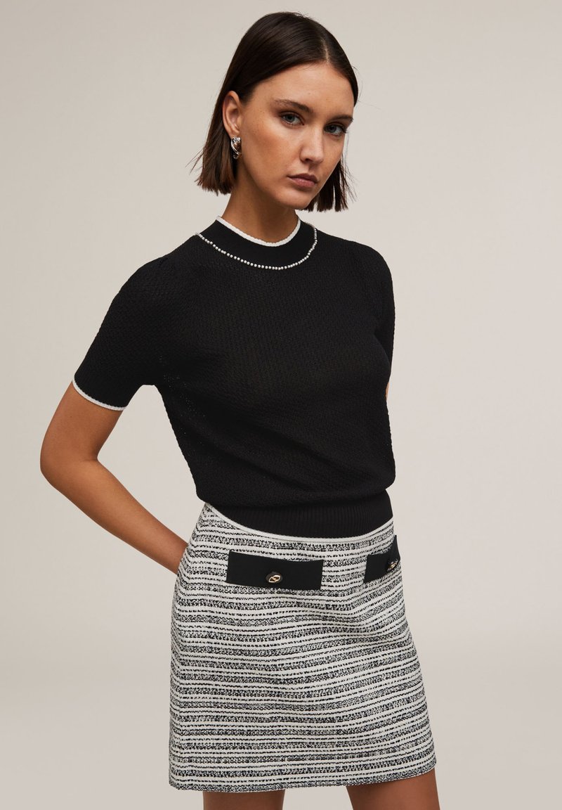 Pull en tricot noir à manches courtes, rehaussé d'une bordure blanche. Associé à une mini-jupe rayée avec des patches noirs et des accents dorés.