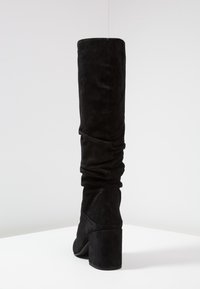 Svart mocka knähög stövel med en slouchad design, chunky klack och mjuk textur. Ingen synlig hårdvara eller detaljer.