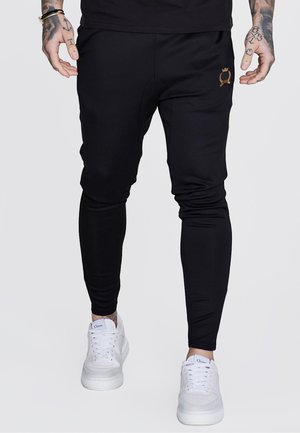Man draagt zwarte slim-fit joggers met gouden embleem en witte sneakers, zichtbaar van taille tot voeten, staand tegen een effen achtergrond.