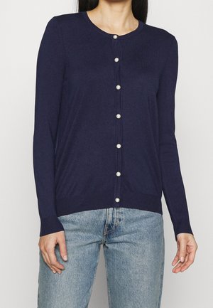 Femme portant un cardigan bleu marine à manches longues avec des boutons en perle sur un jean bleu clair, debout devant un fond clair uni.