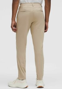 Bărbat îmbrăcat cu pantaloni bej slim-fit și adidași albi, văzut din spate pe un fundal simplu.