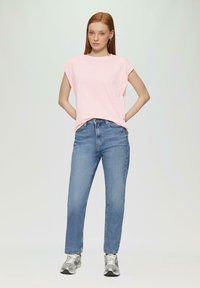 Růžové tričko s krátkým rukávem a ohrnutými okraji, spojené s světle modrými džíny rovného střihu a stříbrnými teniskami. Modelka stojí s rukama na bocích.