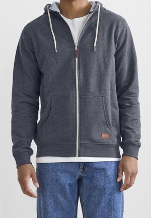 Mężczyzna ubrany w szarą bluzę z kapturem zapinaną na zamek i białym sznurkiem, na białej koszulce i niebieskich dżinsach, stojący na jednolitym tle.