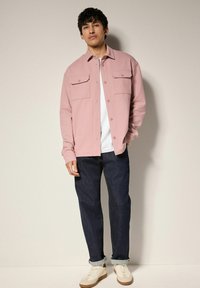 Lys pink button-up skjorte med to brystlommer, båret over en hvid T-shirt. Parret med mørkeblå denim jeans og beige sneakers.