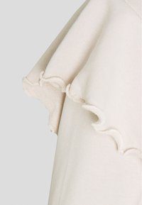 Tejido de algodón de color beige claro con un borde con volantes. La textura es suave, con un tejido sutil y liso. Detalle clave: dobladillo con puntas.